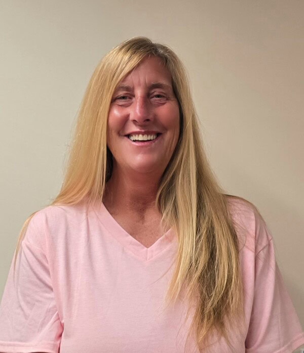 a woman with long blonde hair and a pink v neck shirt smiles while standing in front of a plain beige wall.
