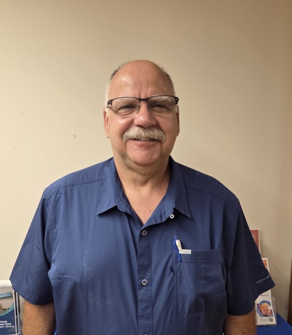 a middle aged man with glasses and a mustache smiles while standing indoors, wearing a blue short sleeve shirt with pens in the pocket.