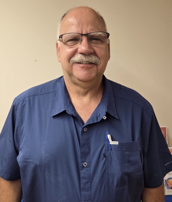a middle aged man with glasses and a mustache smiles while standing indoors, wearing a blue short sleeve shirt with pens in the pocket.