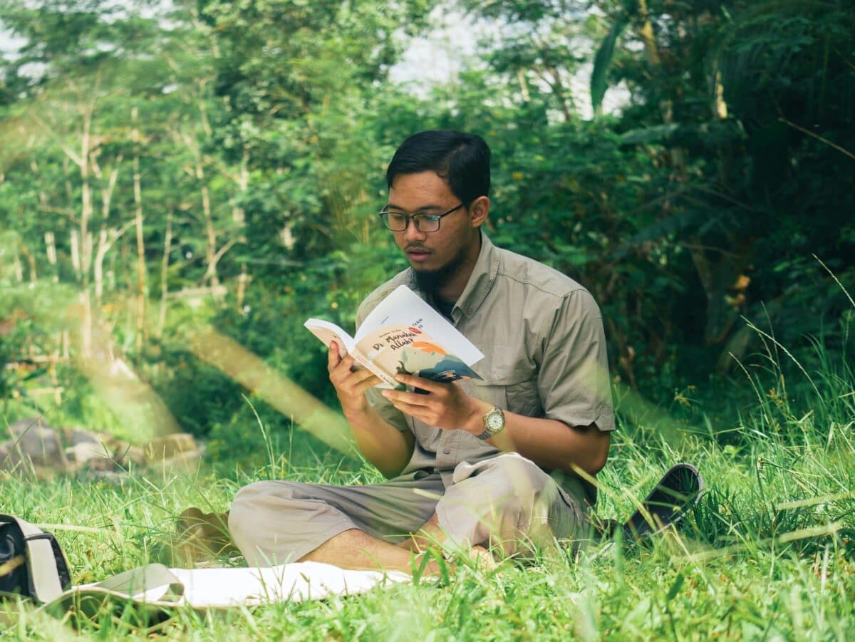 a man sits cross legged on the grass outdoors, reading a book. he wears glasses and casual clothes, with trees and greenery in the background.