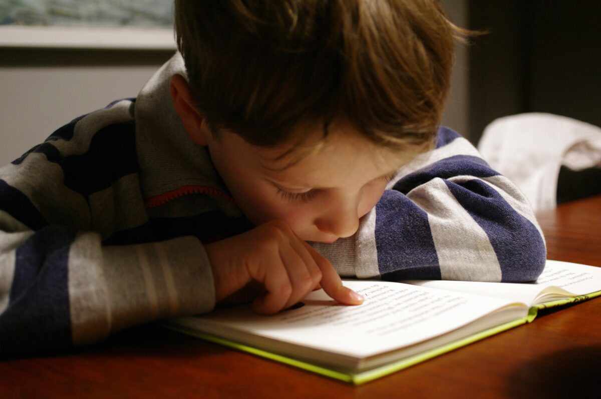 a young boy wearing a striped shirt reads a book at a table, using his finger to follow the text.